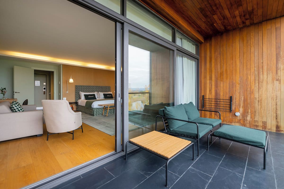 A modern interior with a sofa, chairs, a bed with patterned headboard, and a view through a glass door.