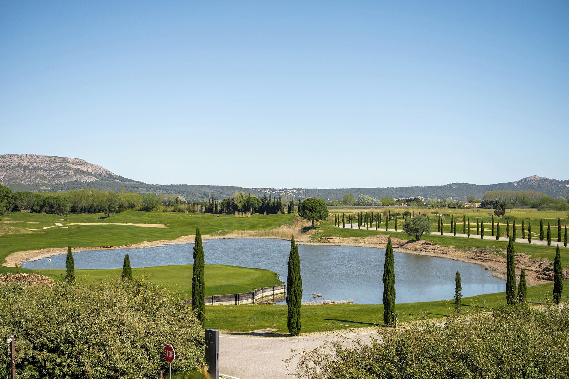 Un campo de golf con dos estanques, cipreses y una montaña al fondo.