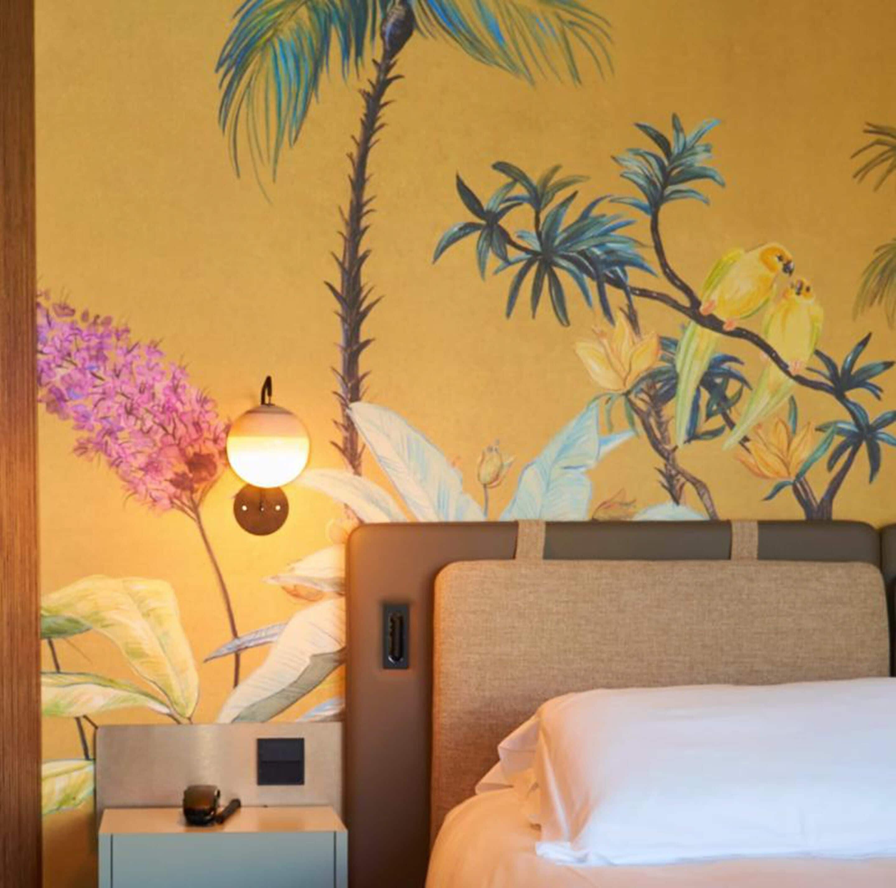 A hotel room with a bed, a nightstand with a lamp, and a wall mural of tropical birds and plants.