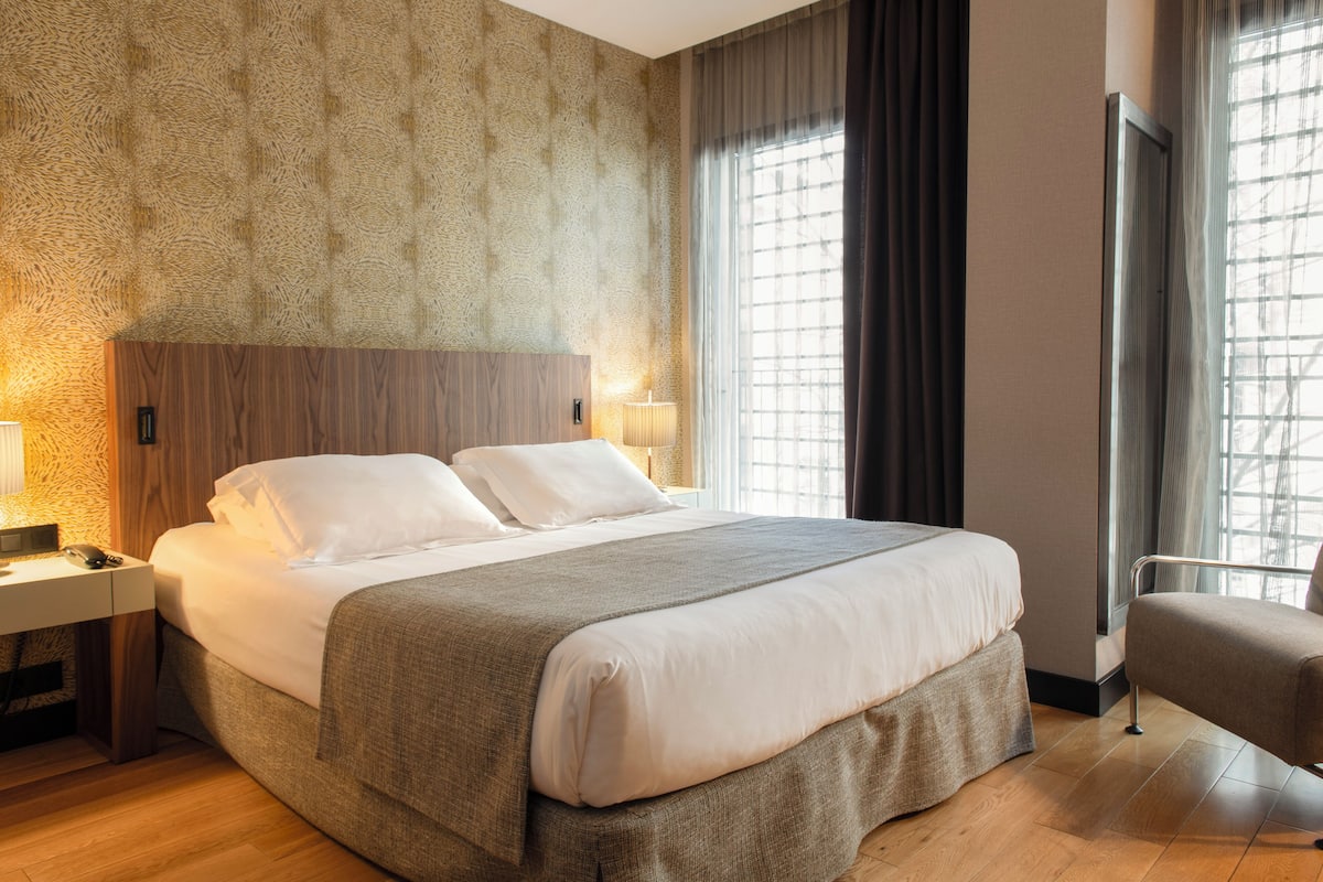 Un lit bien fait, avec du linge de lit blanc et un plaid gris, une table de chevet en bois avec une lampe, et une fenêtre avec des rideaux.