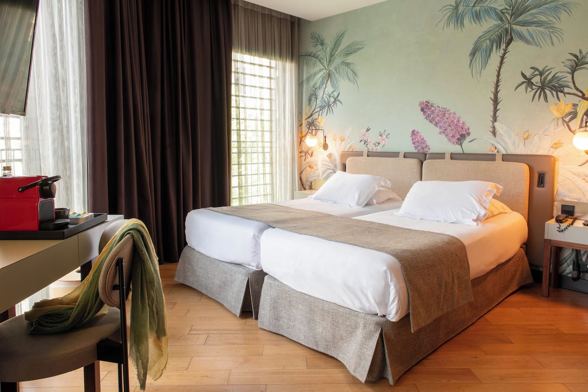 Un lit double avec une tête de lit beige, un bureau avec une chaise rouge et un mur orné d’une fresque aux motifs tropicaux.