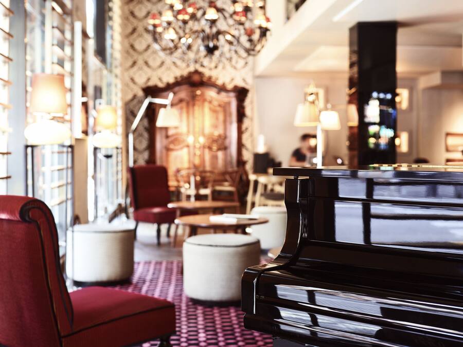 A grand piano in a well-lit, elegantly decorated room with a patterned carpet, plush seating, and a chandelier.