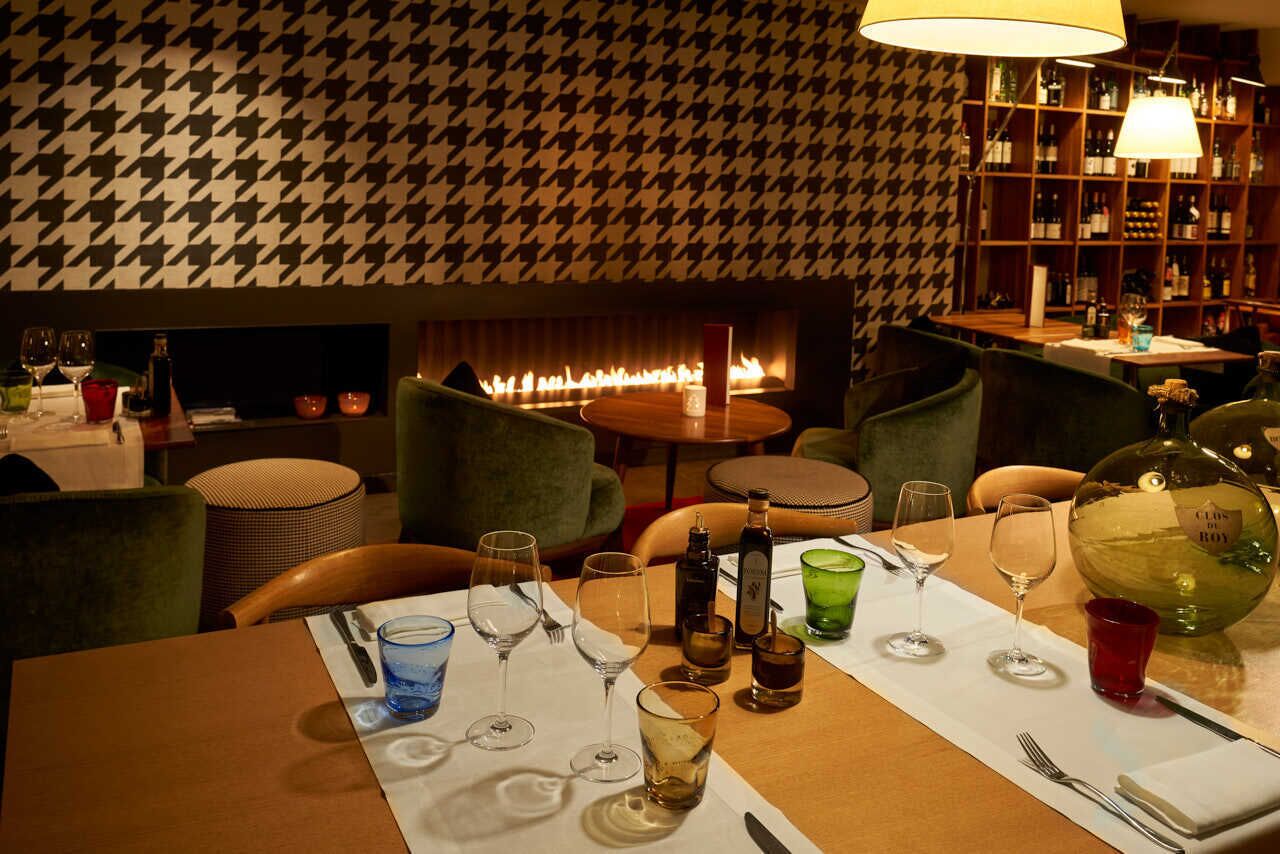 A cozy restaurant with a houndstooth patterned wall, a fireplace, and a bar area with shelves of bottles.