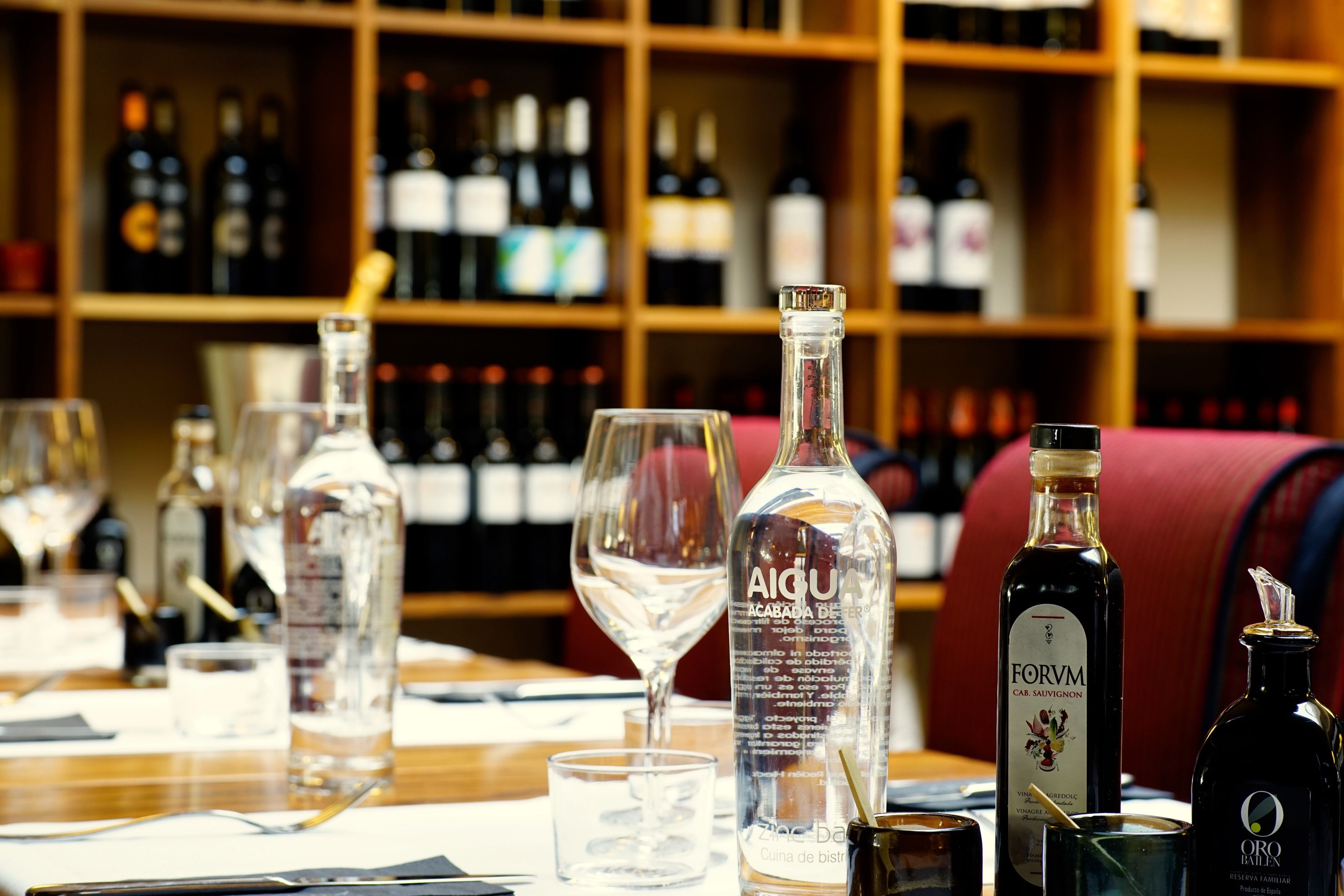 Een barscène met diverse flessen en glazen, waaronder een opvallende fles met de aanduiding 'AIGU' en een andere met 'FORVM'.