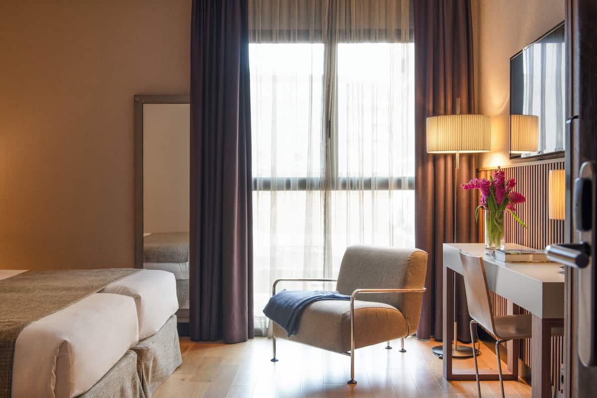 Une chambre d’hôtel avec un lit, un bureau, une chaise, une lampe et une fenêtre avec des rideaux.