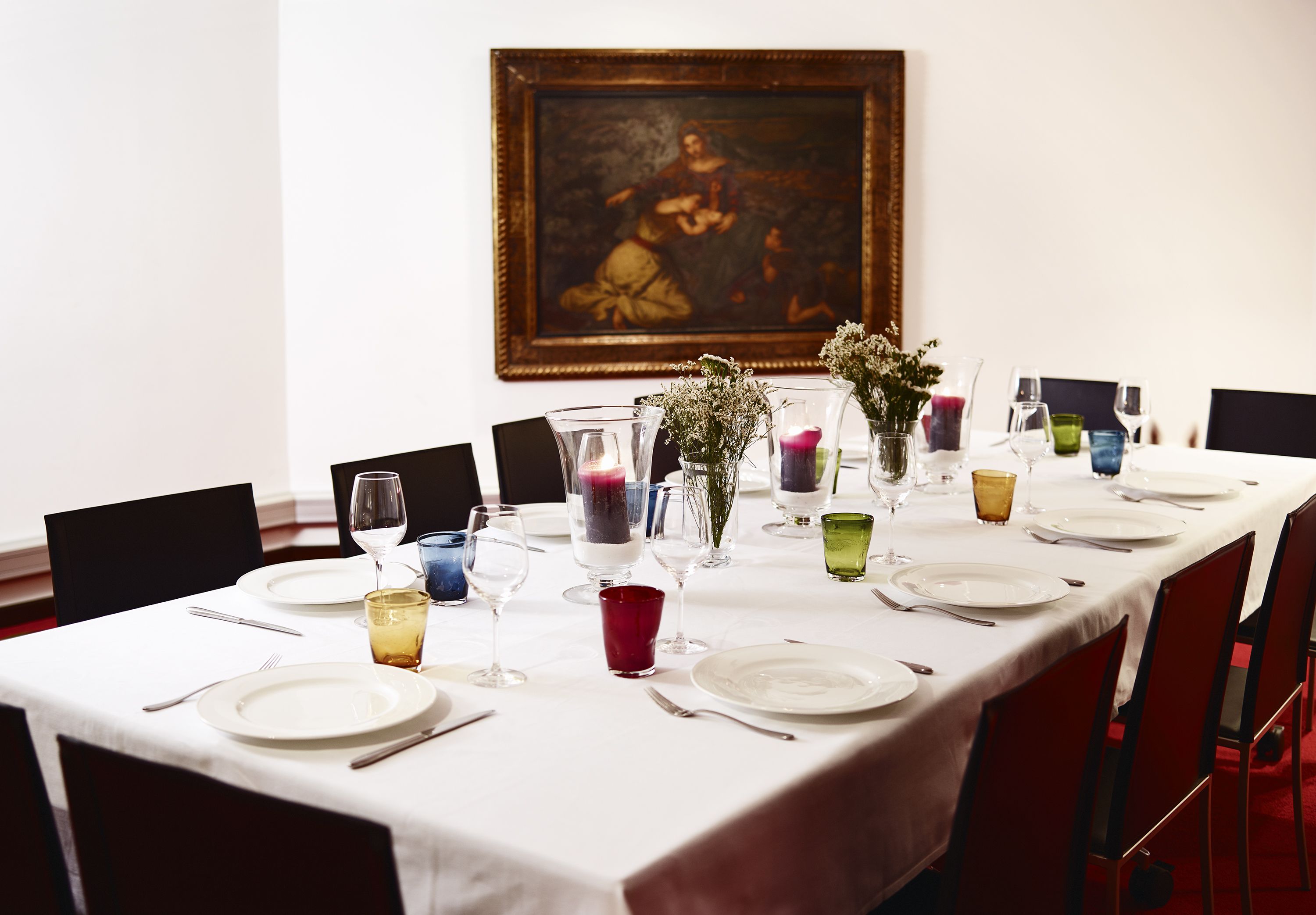 A dining table set for a meal with a white tablecloth, clear glassware, and a framed painting on the wall.