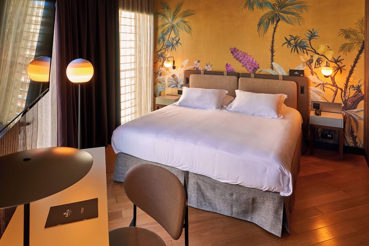 Une chambre d’hôtel avec un grand lit, un bureau et une chaise. On y trouve deux tables de chevet avec des lampes, ainsi qu’une fresque murale représentant des plantes tropicales.