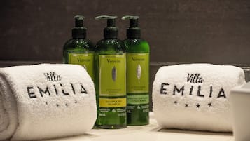 Trois bouteilles de shampoing et d’après-shampoing à la verveine, et deux serviettes sur lesquelles est brodé le nom « Villa Emilia », posées sur le comptoir d’une salle de bain.