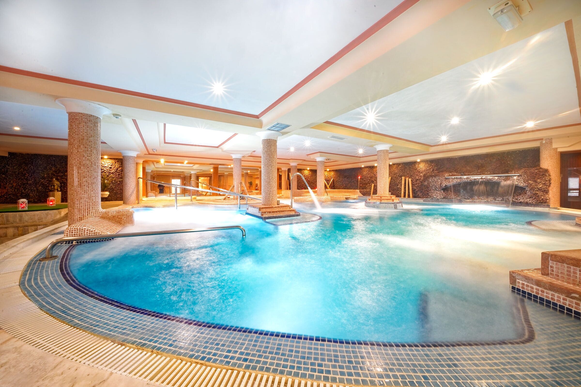 A large indoor swimming pool with multiple hot tubs, a stone wall, and a tiled floor.