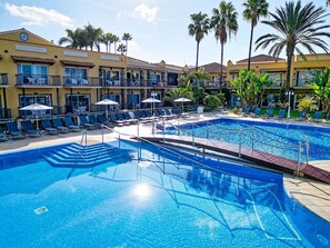 A swimming pool area with lounge chairs and umbrellas, surrounded by a building with balconies and palm trees. - Bull Vital Suites & Spa Boutique Hotel - Only Adults (San Bartolomé de Tirajana)