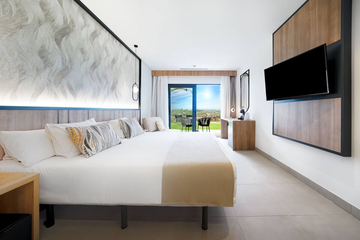 A modern hotel room with a large bed, a flat-screen TV mounted on the wall, and a balcony with a view of the city.