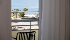 Vista desde una ventana con una silla al frente, que da a una zona costera con carretera, coches aparcados y vegetación.
