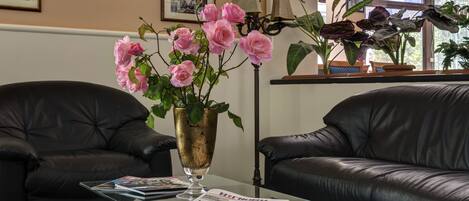 Um quarto com dois sofás de couro preto, uma mesa de centro de vidro com revistas, um vaso com rosas cor-de-rosa e uma janela grande com plantas.