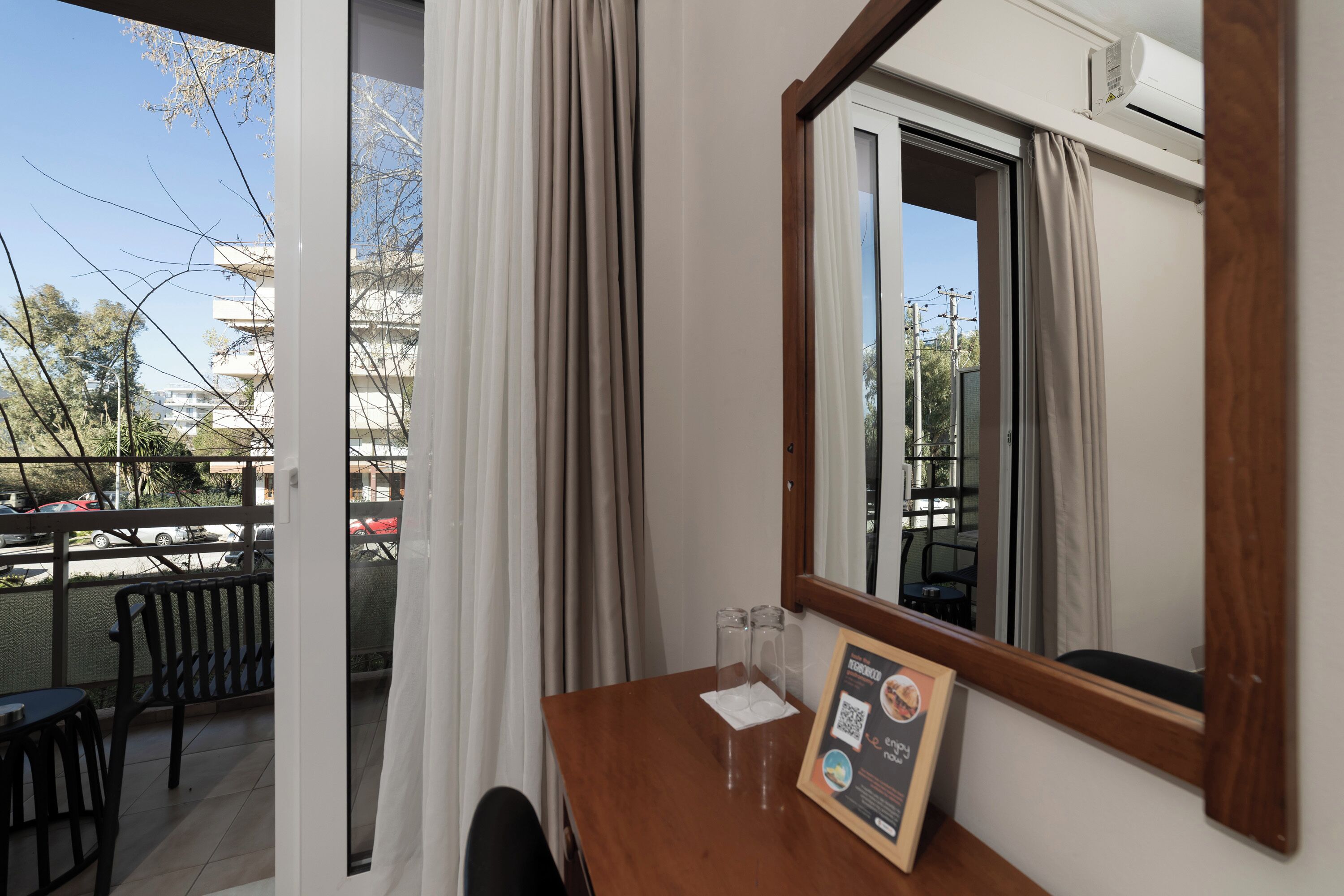 A balcony with a view of the city, a wooden table with a framed menu, and a window with curtains.