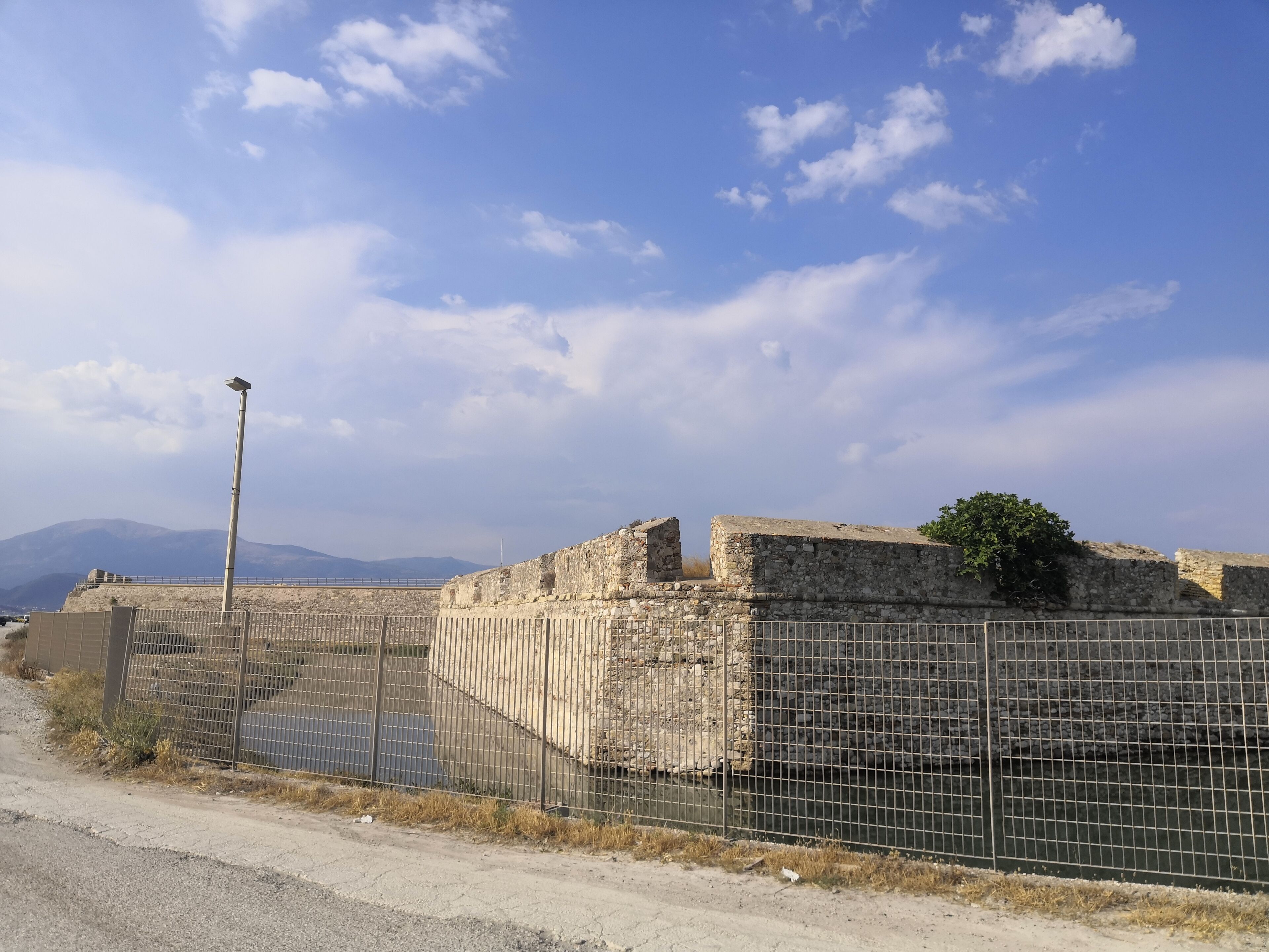Eine historische Steinmauer mit einem Metallzaun im Vordergrund, eine Straßenlaterne und ein klarer Himmel.