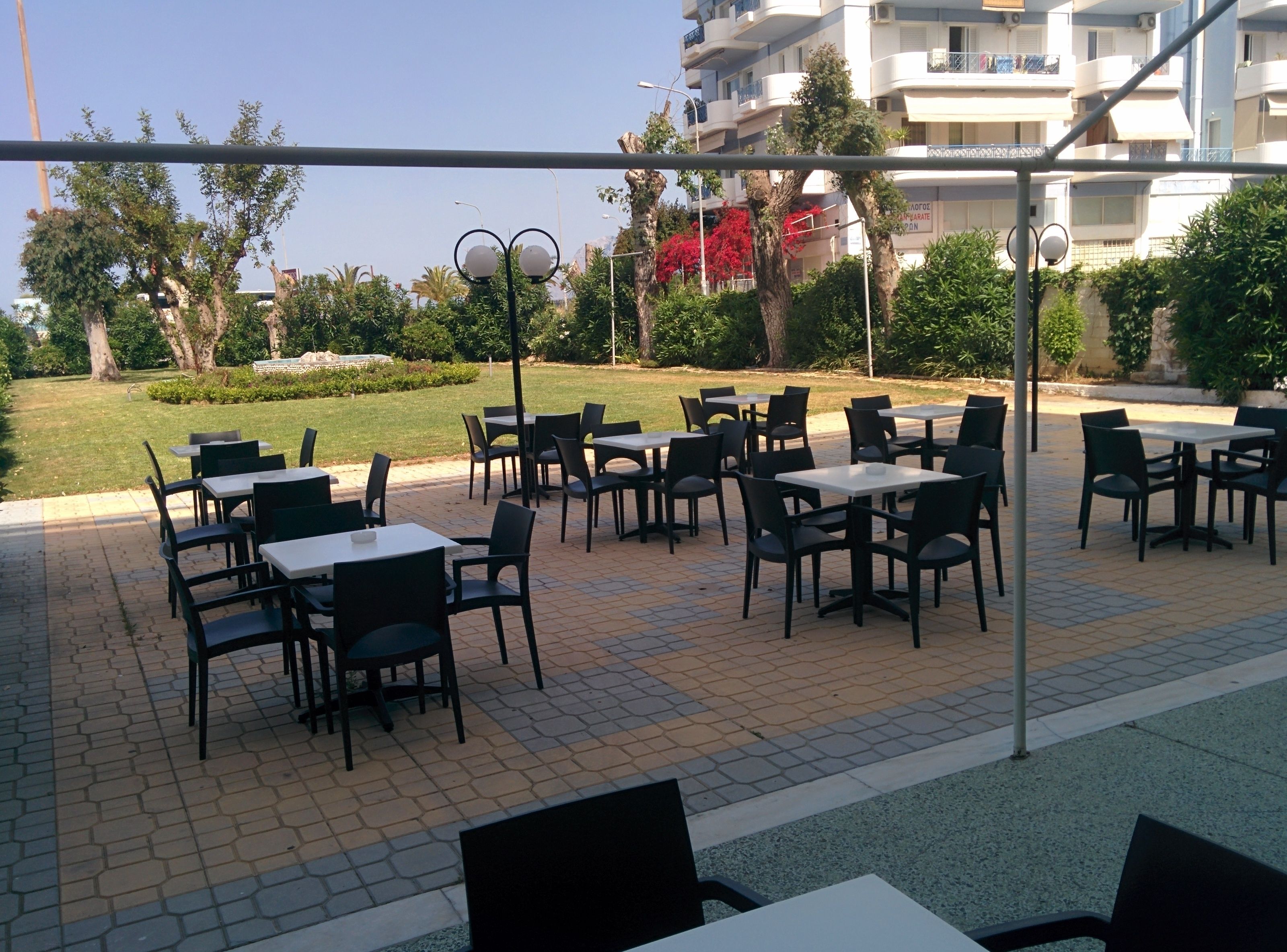 An outdoor seating area with tables and chairs, surrounded by greenery and a building in the background.