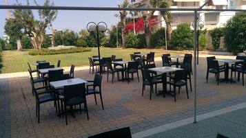 An outdoor seating area with tables and chairs, surrounded by greenery and a building in the background.