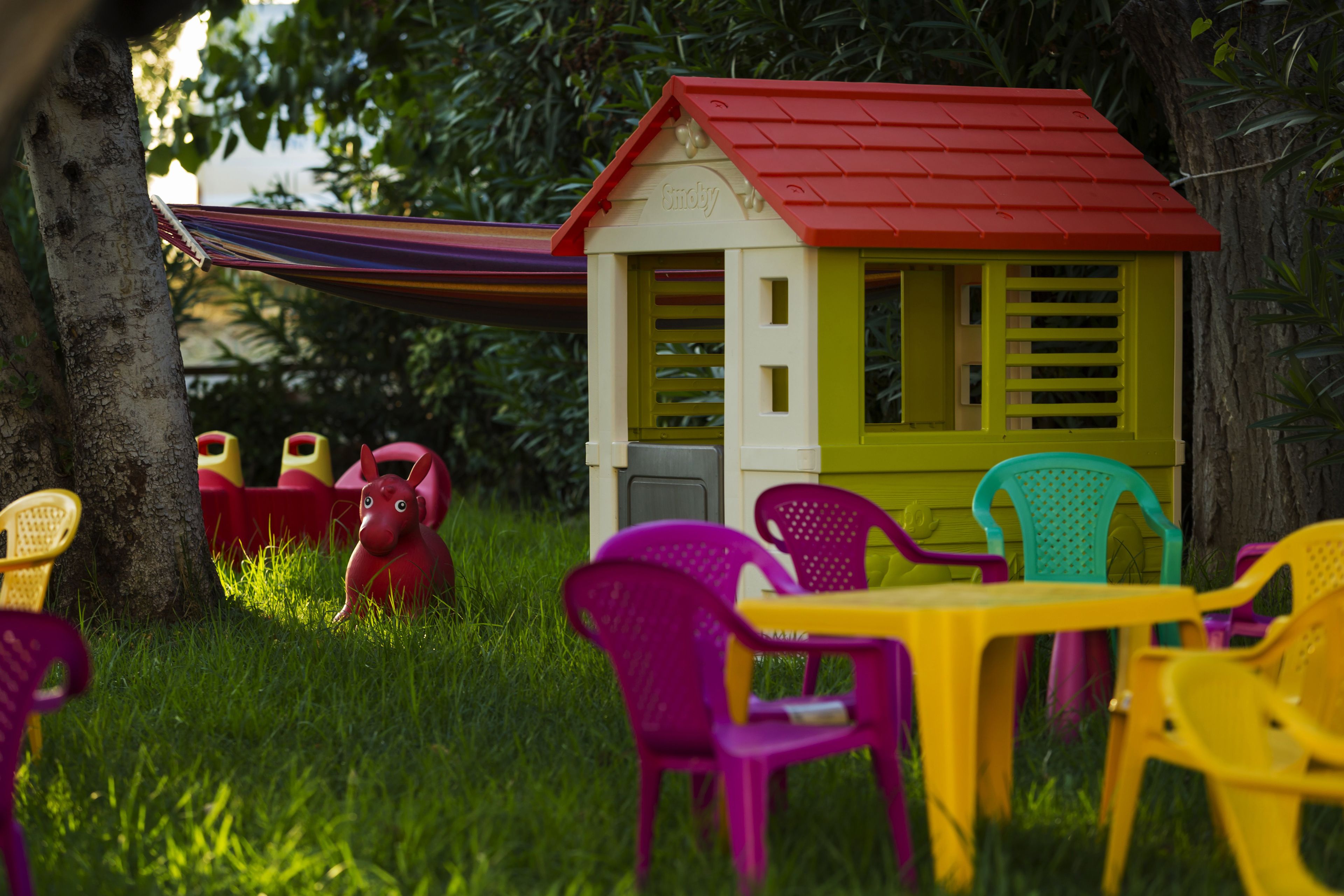 A small playhouse with a red roof and yellow walls, surrounded by colorful plastic chairs and a hammock.