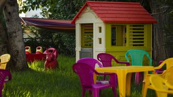 A small playhouse with a red roof and yellow walls, surrounded by colorful plastic chairs and a hammock.