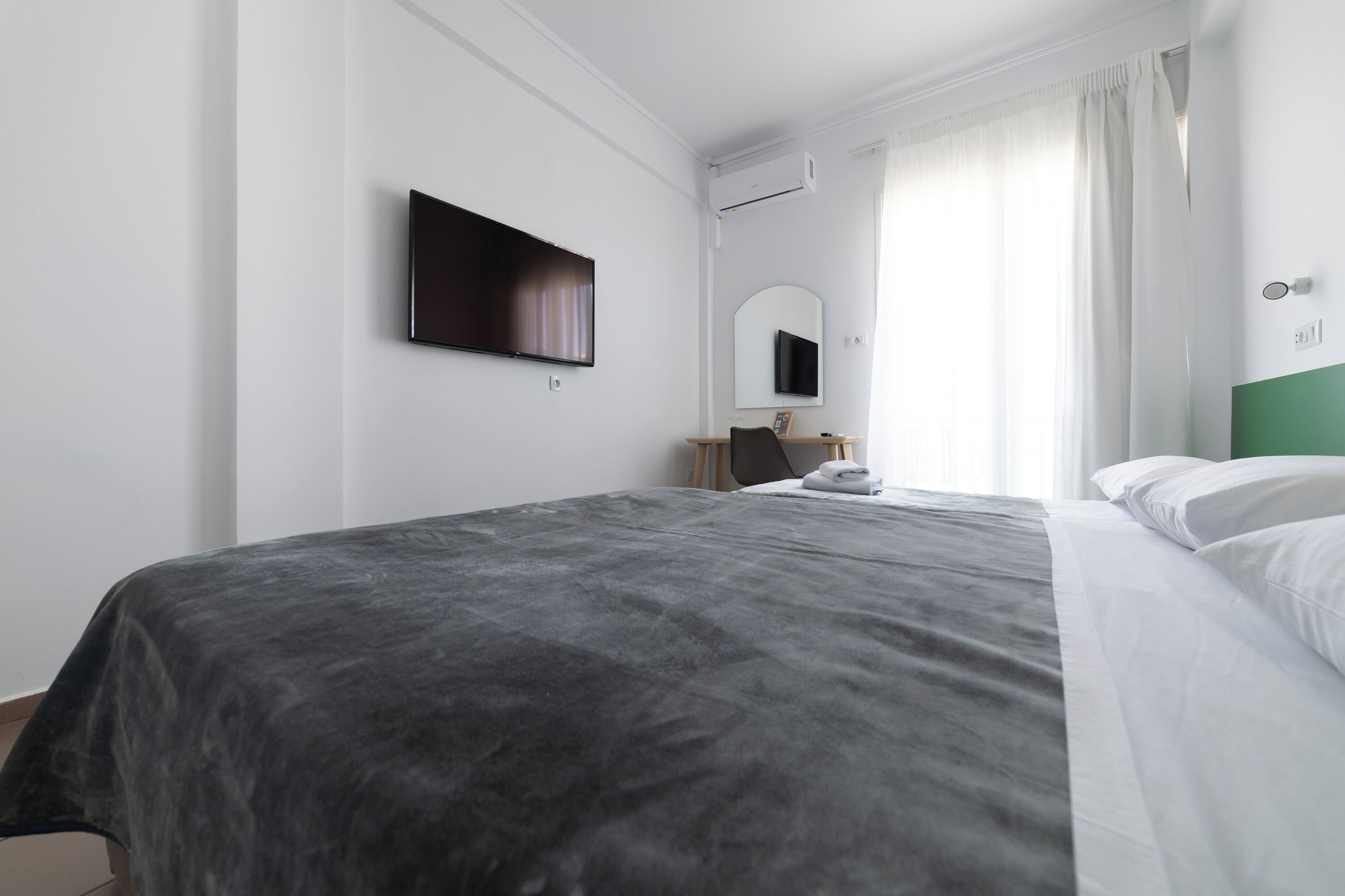 Una habitación de hotel con cama, televisor de pantalla plana en la pared, espejo y ventana con cortinas.