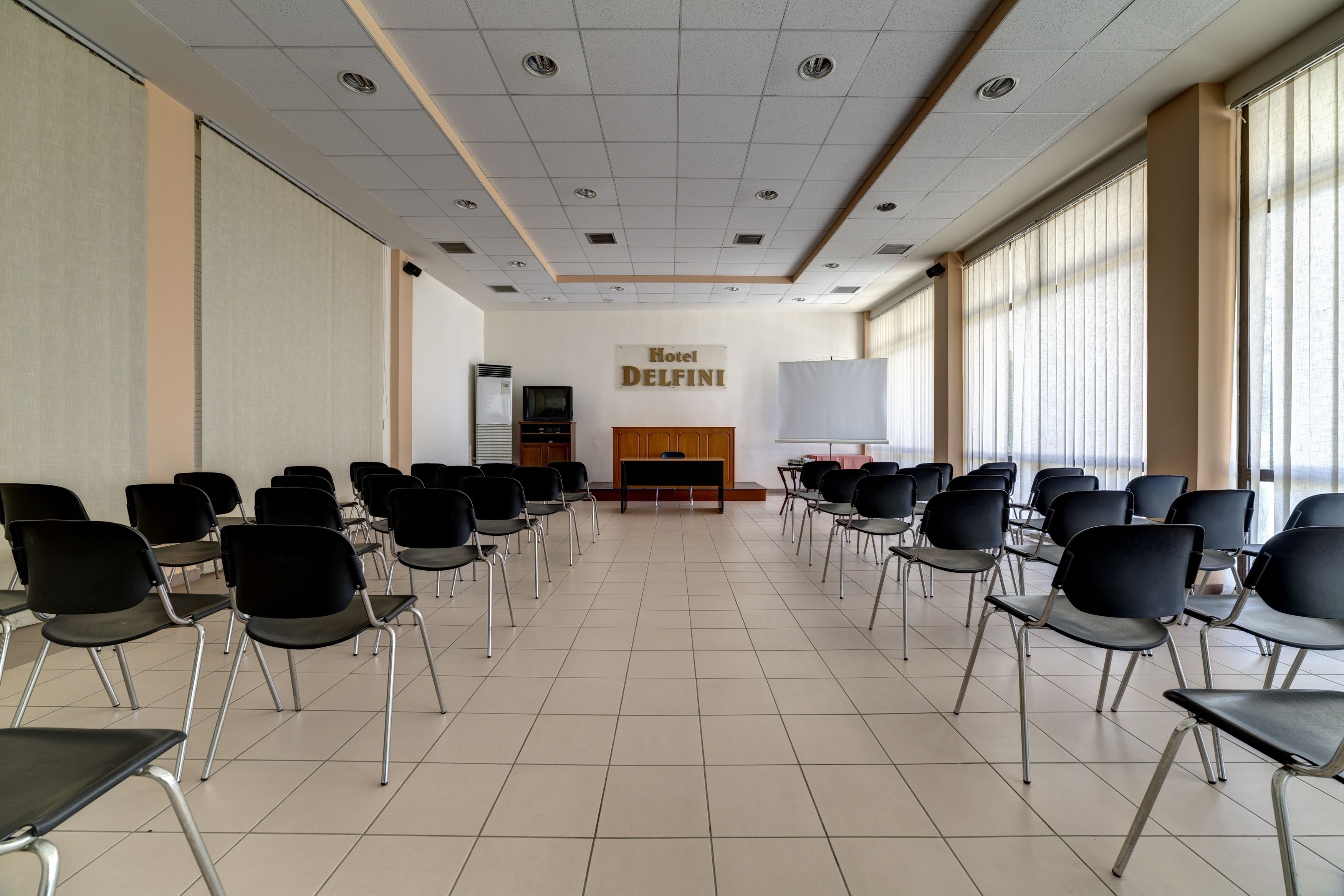 Un salón de conferencias con filas de sillas, un atril y un cartel que dice 'Hotel Delfini'.