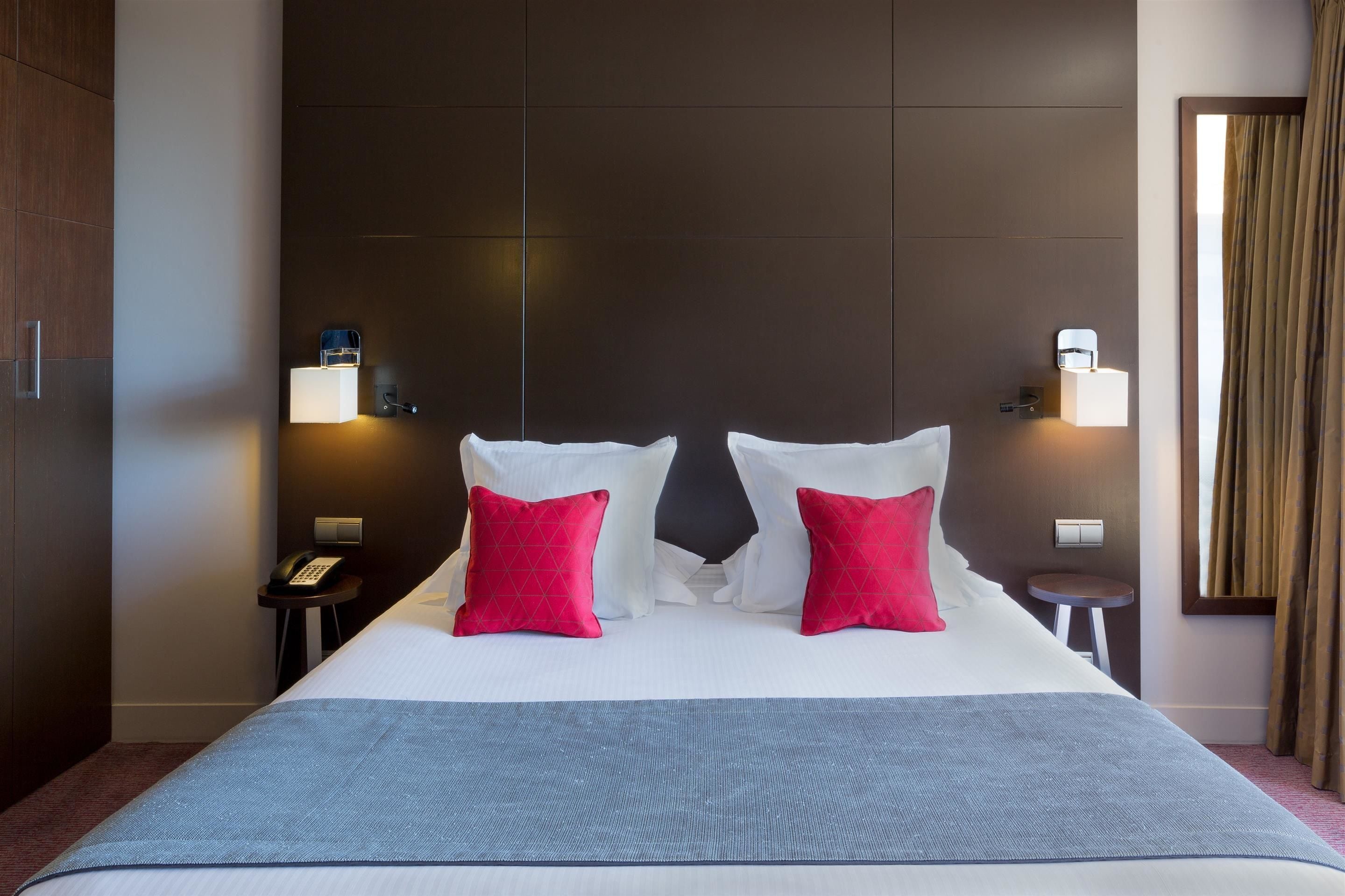 A hotel room with a large bed, two red pillows, a blue blanket, a nightstand with a phone, and a wall-mounted lamp.
