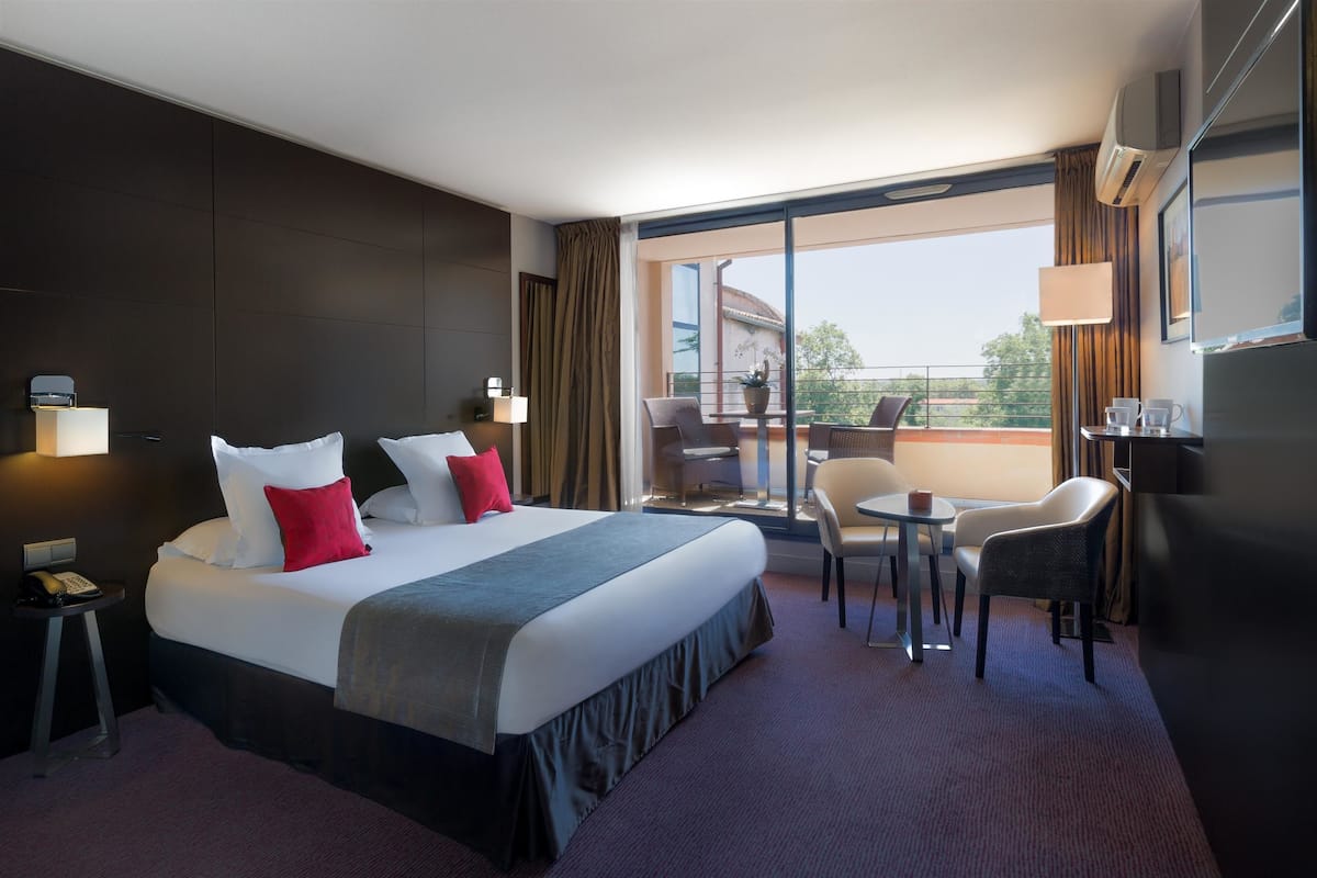 Quarto de hotel com cama grande, varanda com mesa e cadeiras e escrivaninha com luminária.