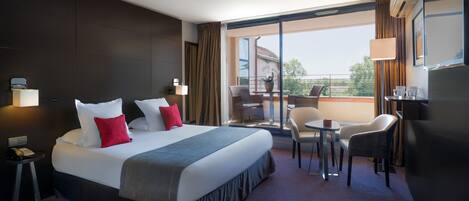 Habitación de hotel con una cama grande, un balcón con mesa y sillas, y un escritorio con lámpara.