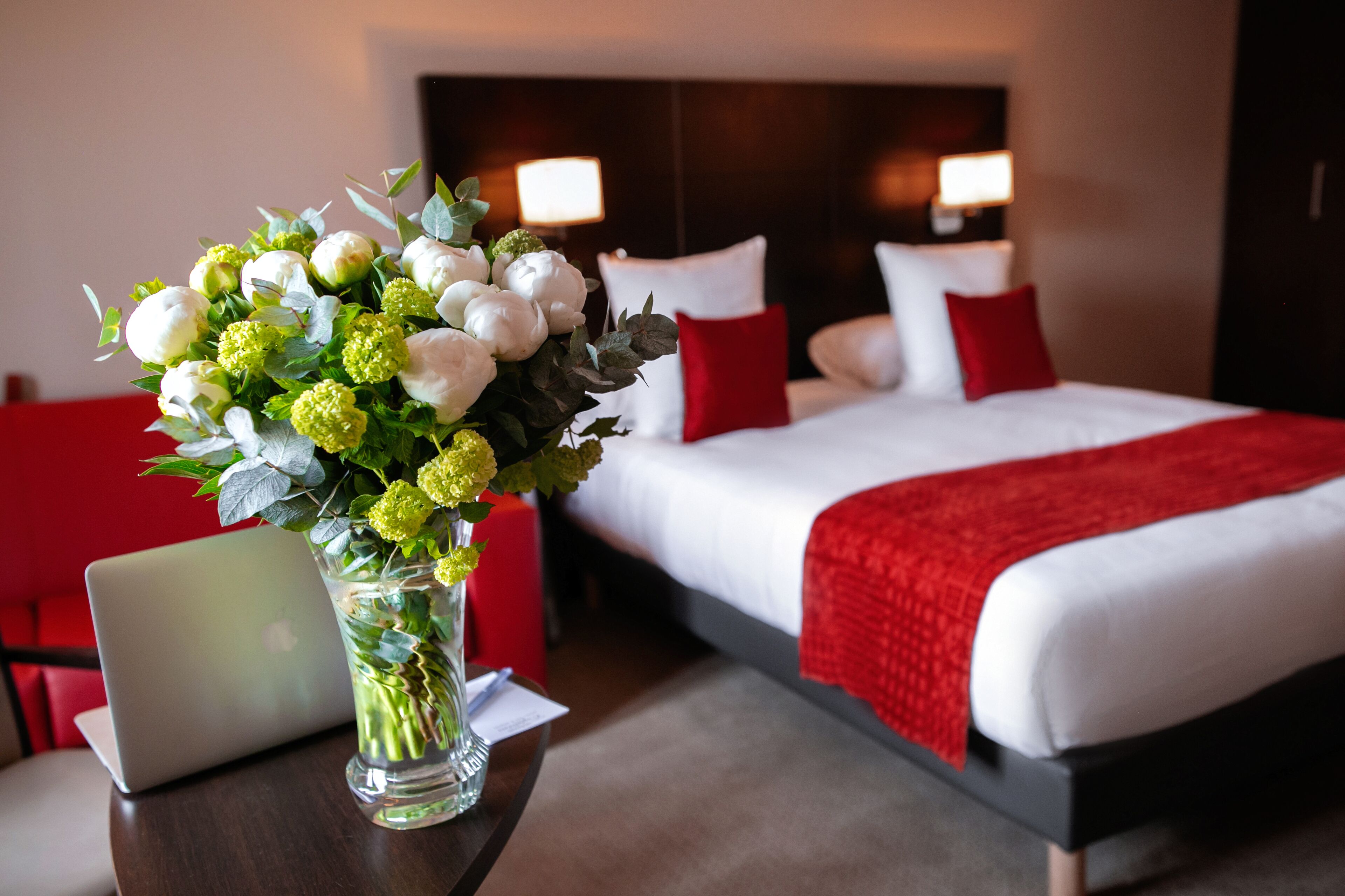Ein Hotelzimmer mit Bett, roten Kissen, einer roten Decke, einem Laptop auf einem Tisch und einer Vase mit Blumen.