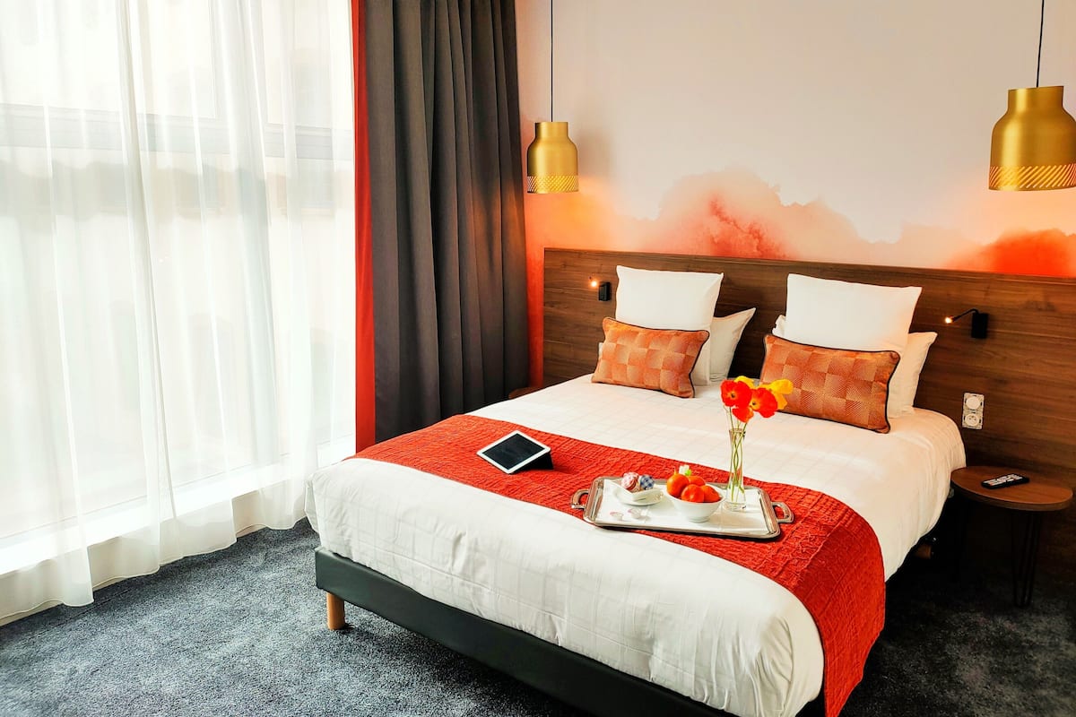 Quarto de hotel com uma cama grande, mesa de cabeceira, luminária na parede e uma janela com cortinas leves.