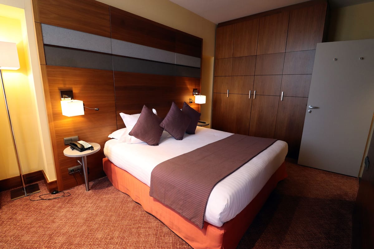 Quarto de hotel com uma cama grande, duas luminárias de cabeceira, uma mesinha com um telefone e um guarda-roupa.