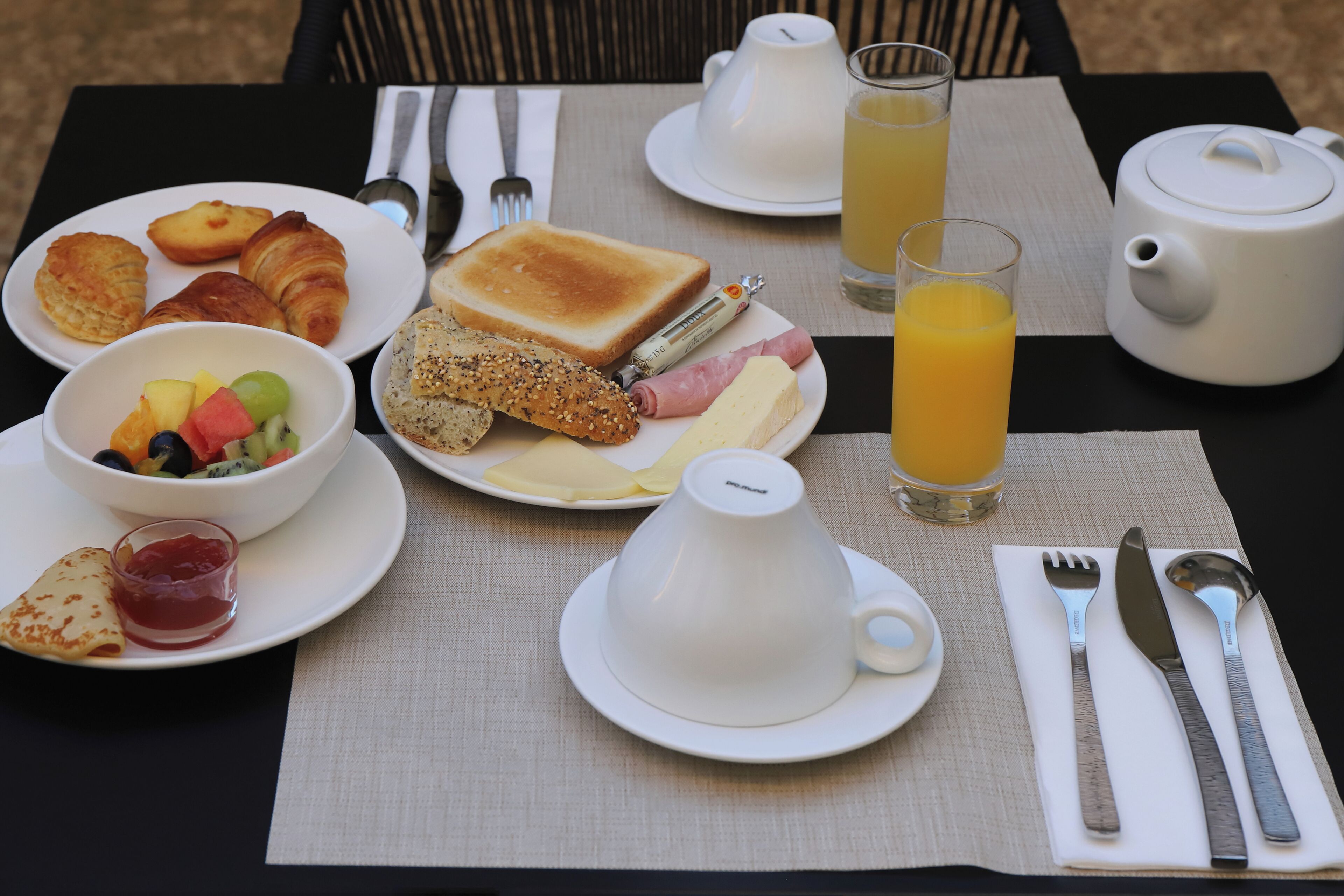 A breakfast setting with croissants, toast, ham, cheese, and fruit on plates, a bowl of mixed fruit, a cup of coffee, and glasses of orange juice.