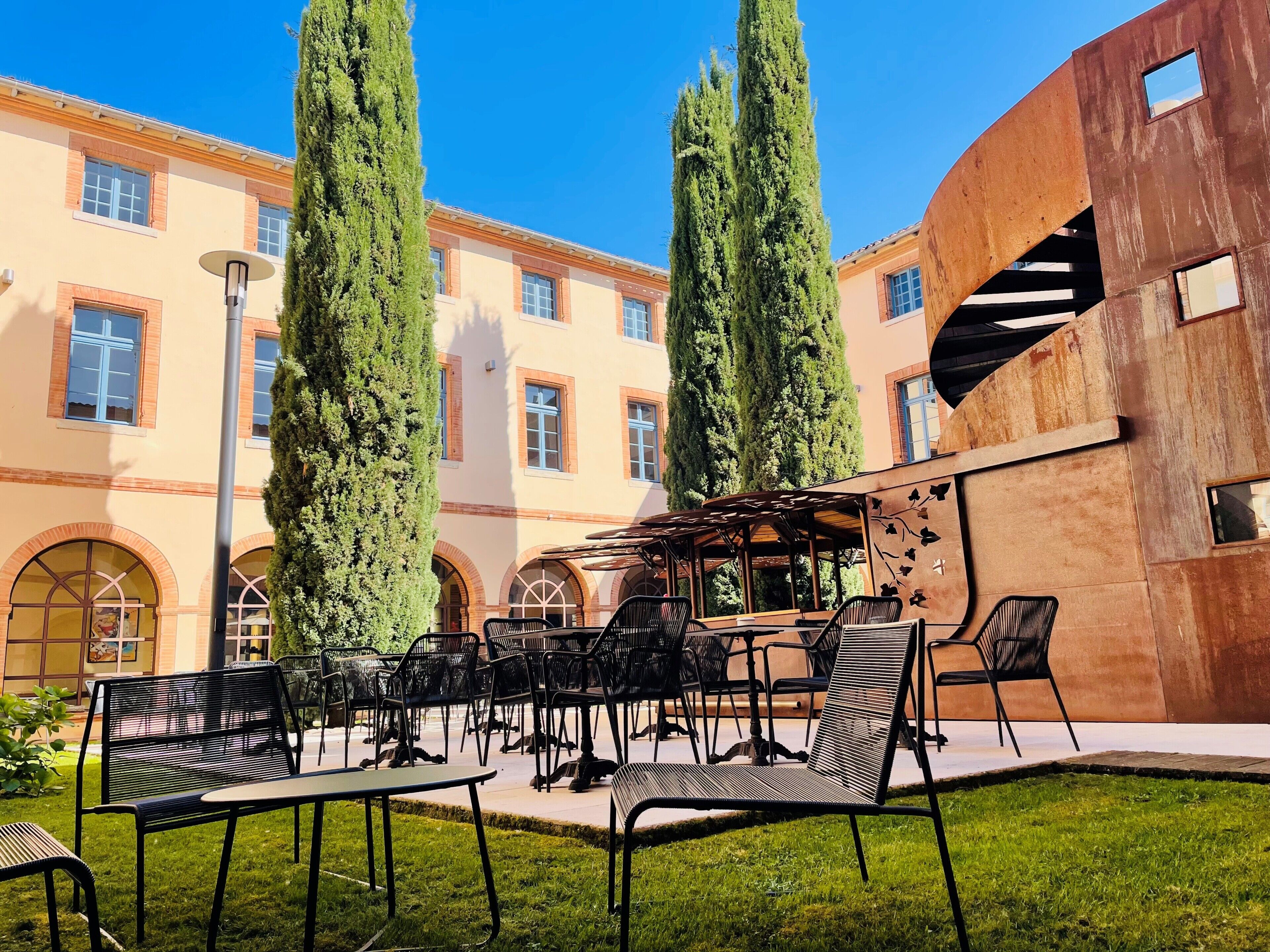 An outdoor seating area with tables and chairs, surrounded by a building with arched windows and a courtyard.