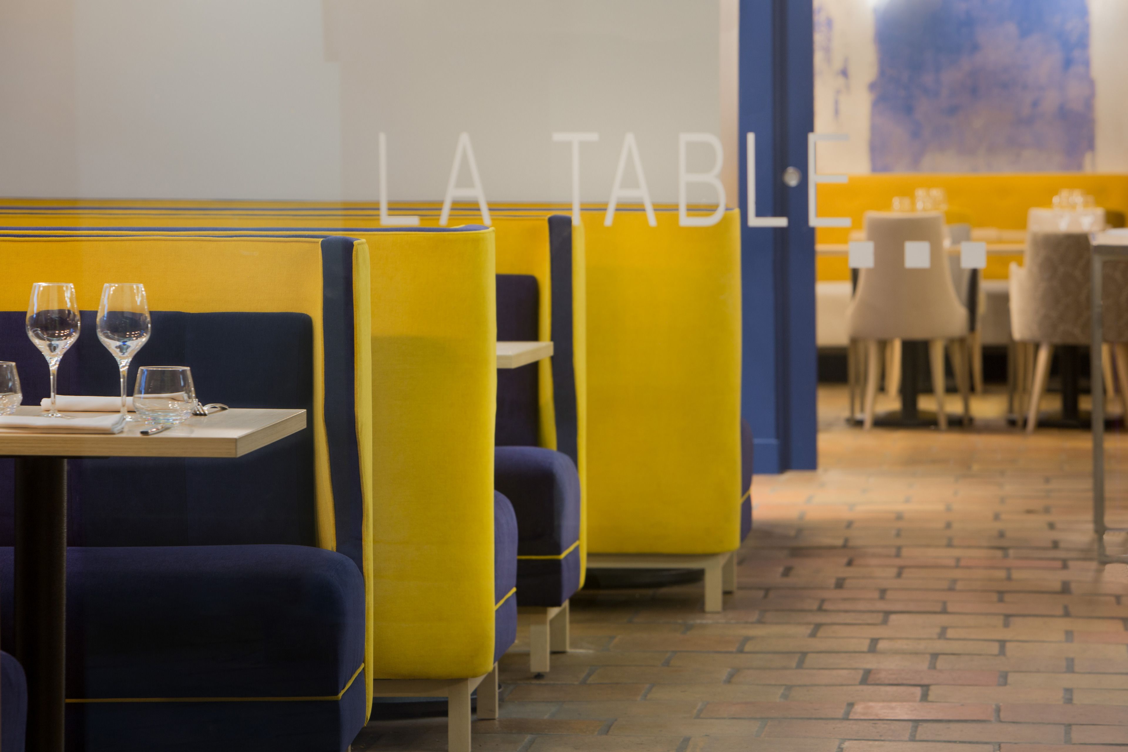 A restaurant interior with yellow and blue booths, tables with glasses and cutlery, and a tiled floor.