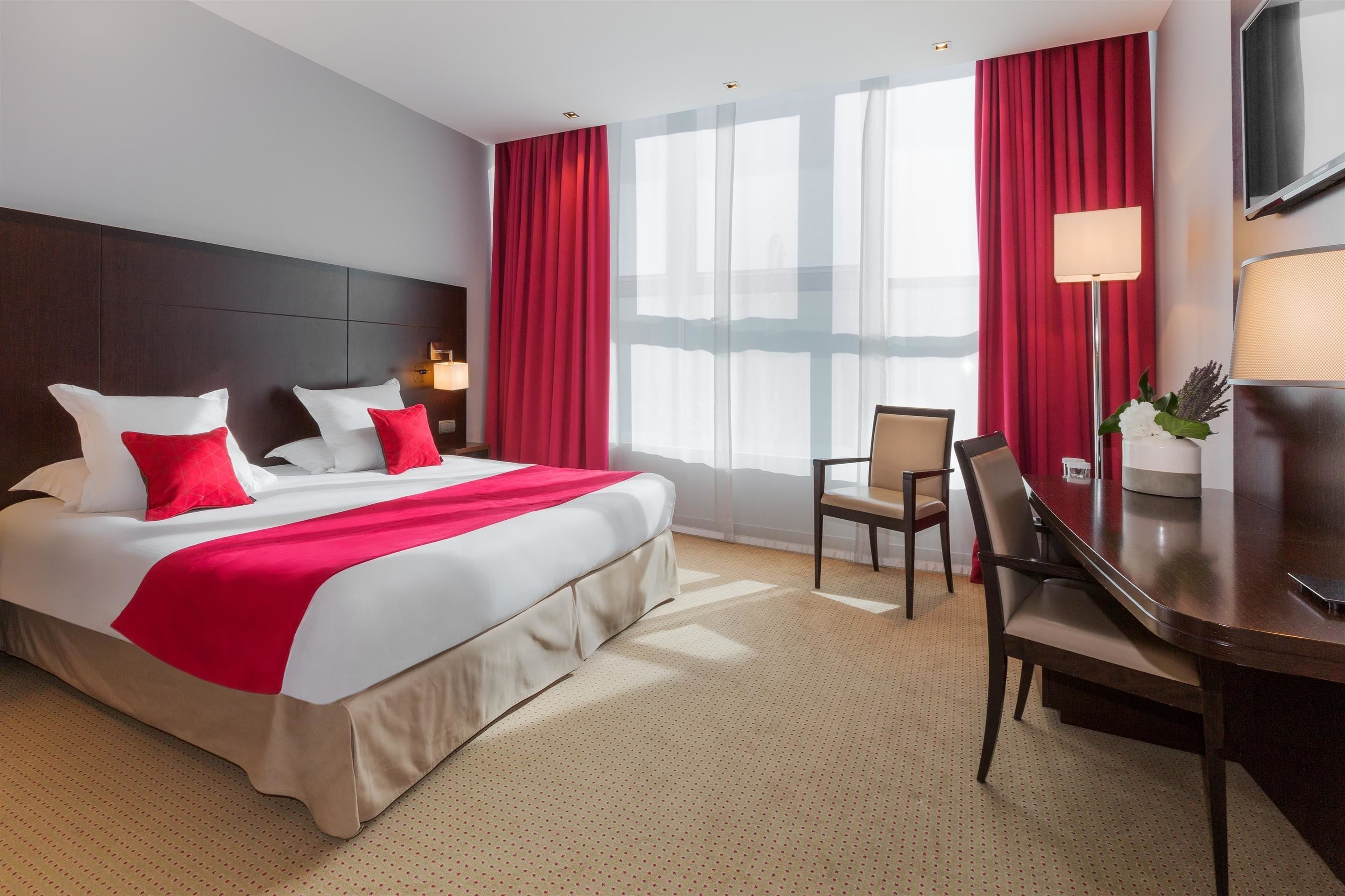 A hotel room with a large bed, red curtains, a desk with a chair, a TV, and a lamp.