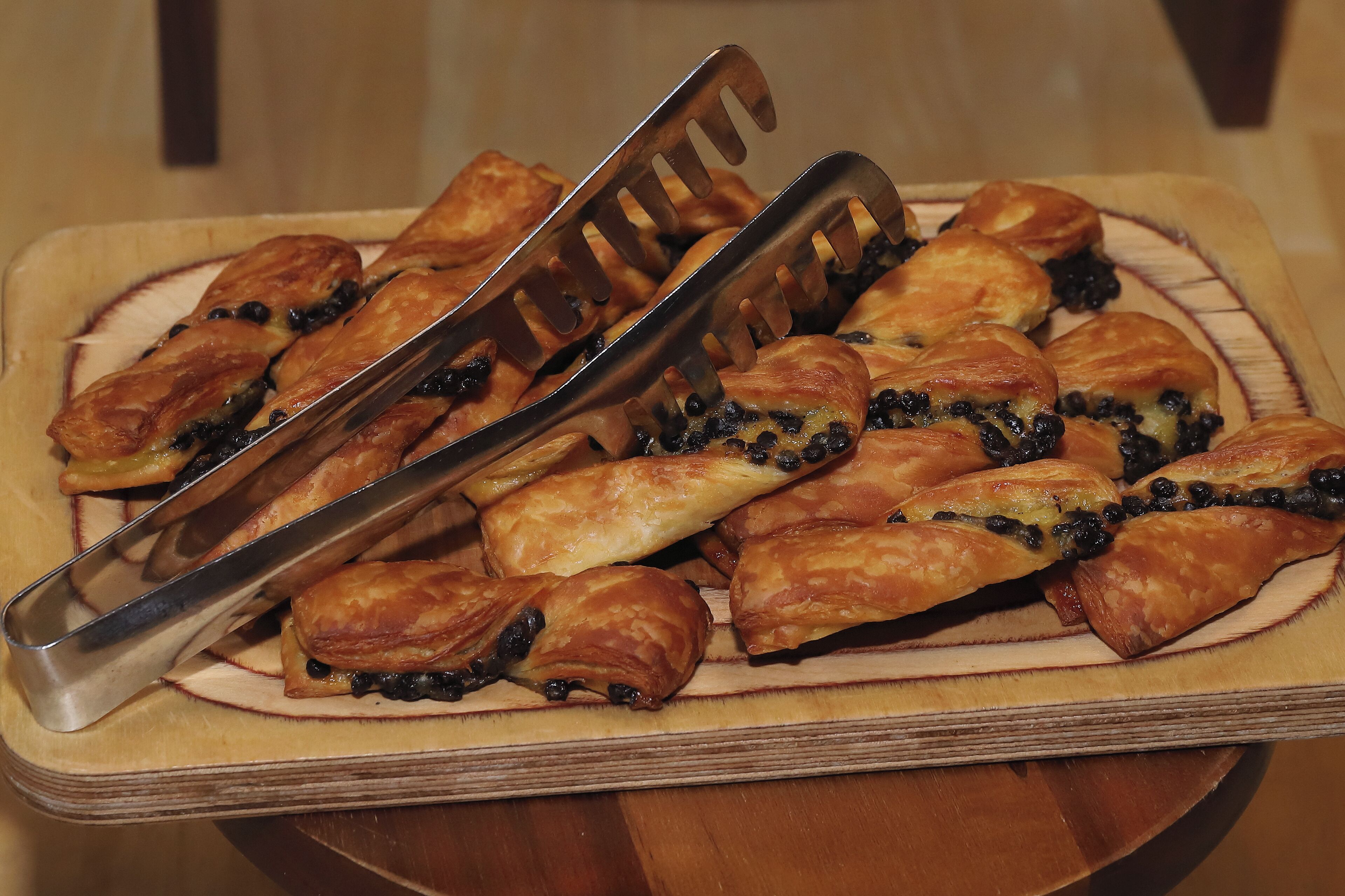 Una tabla de madera con una pila de pasteles dorados, algunos con semillas negras visibles, y un par de pinzas metálicas encima.
