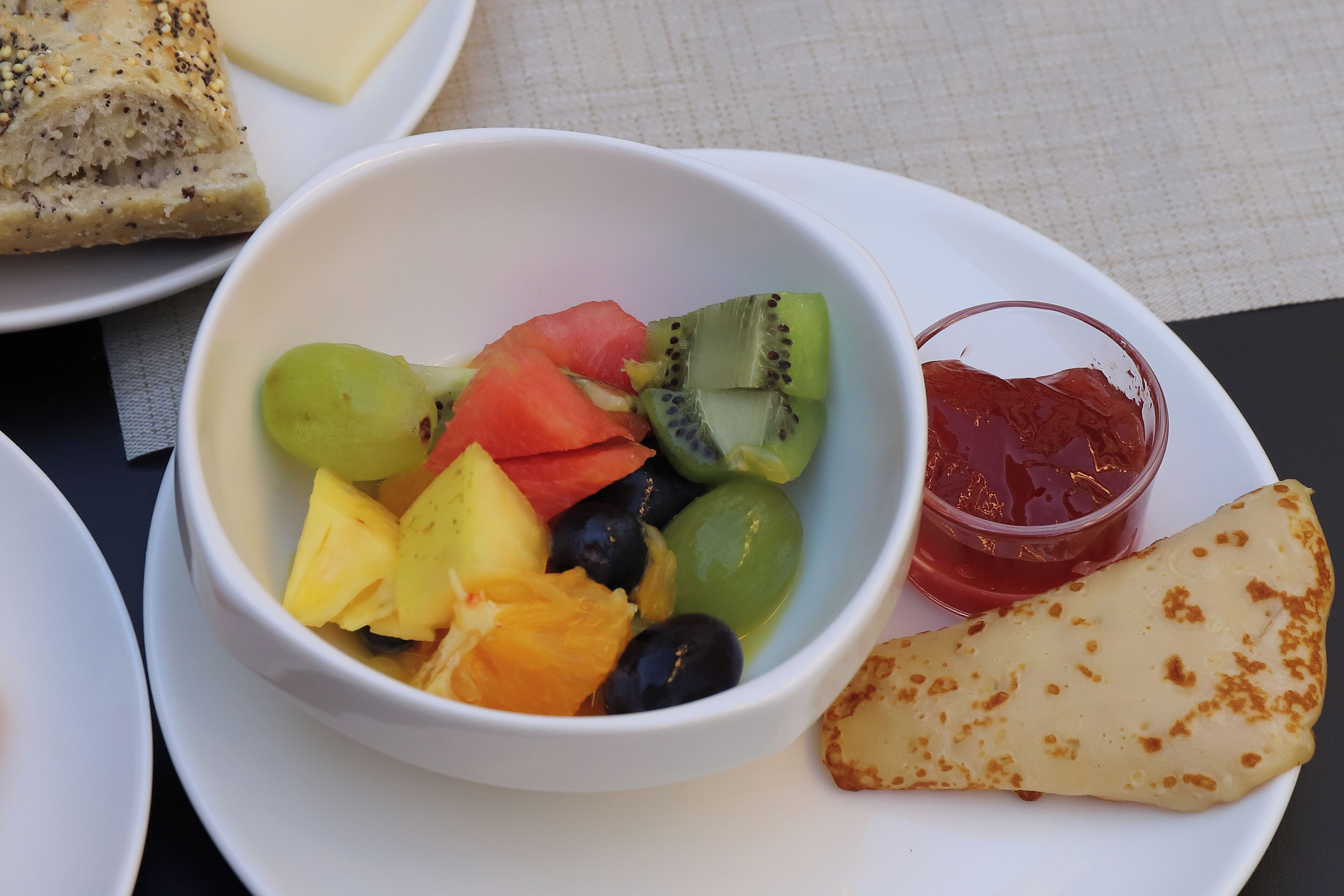 A meal with a bowl of fruit, a slice of cheese, and a small container of jam on a plate.