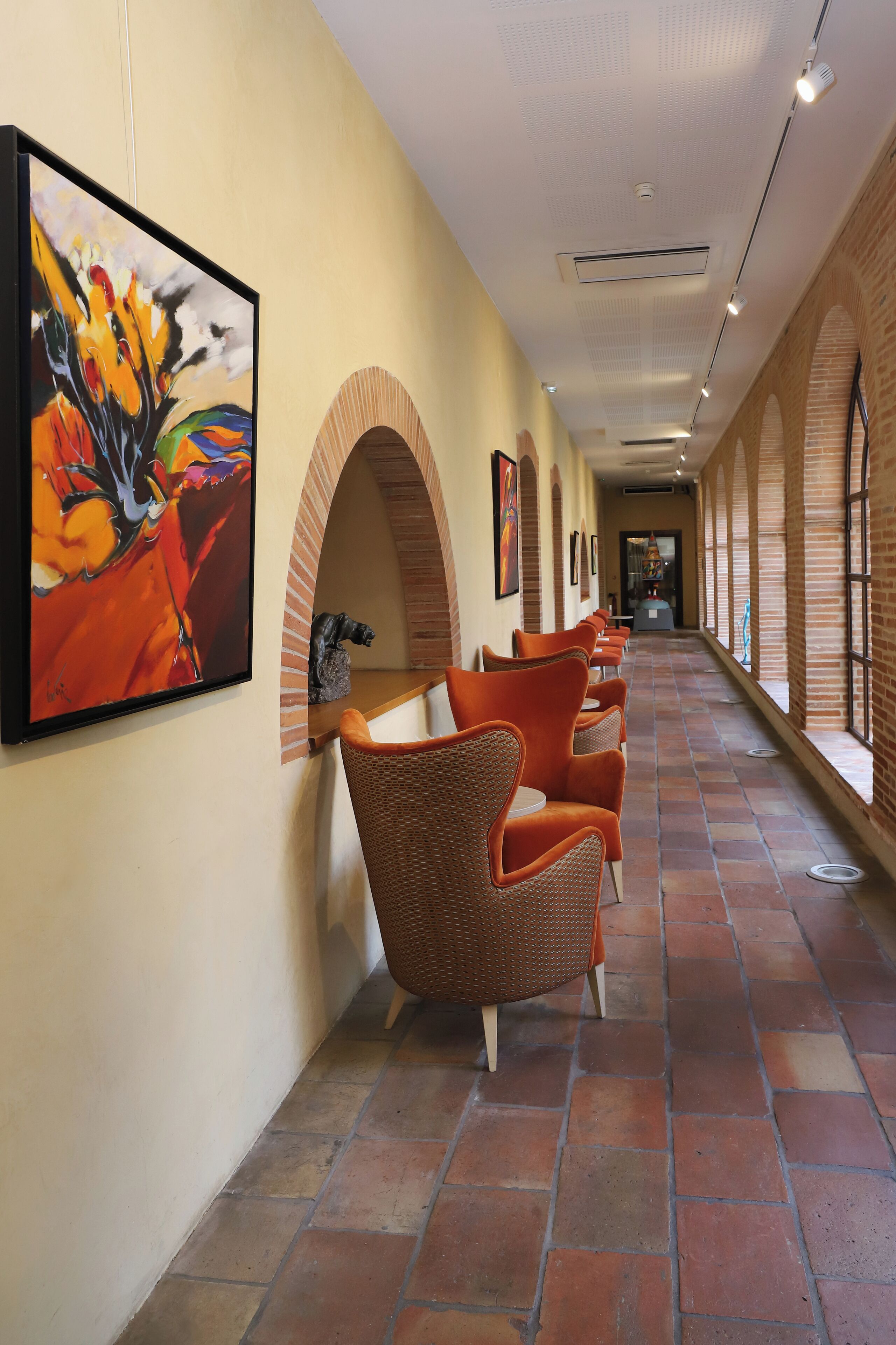 A corridor with brick arches, orange chairs, and a painting on the wall.