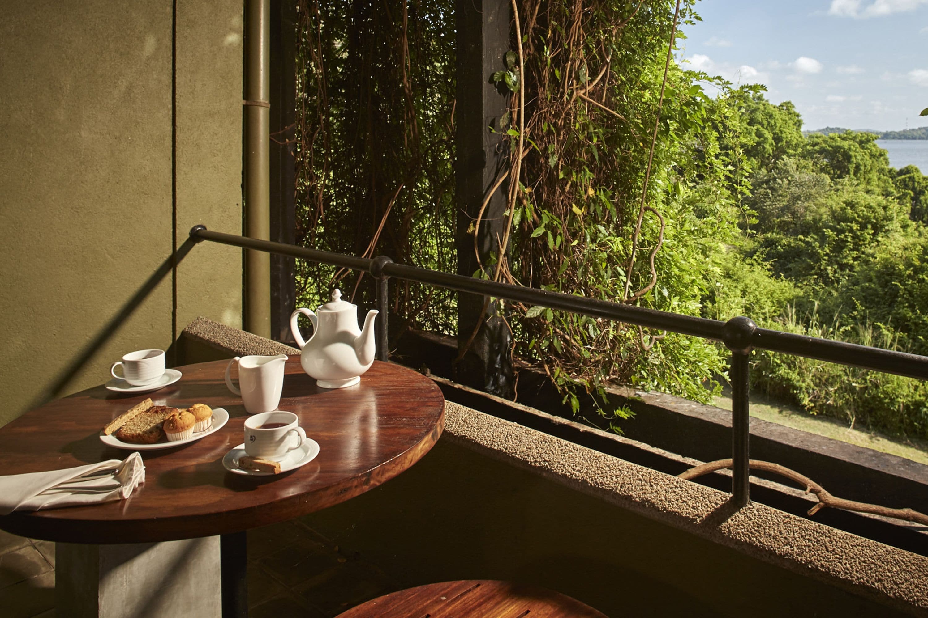 A wooden table with a white teapot, cups, and pastries on a balcony overlooking a lush green landscape and a body of water.