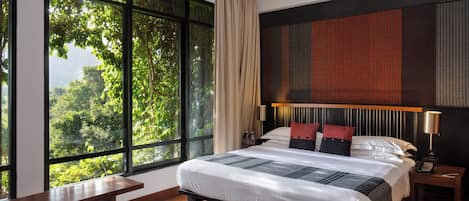 A modern hotel room with a large bed, a wooden bench, a nightstand, and a view of lush greenery through large windows.