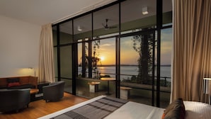 A modern hotel room with a large bed, a sofa, and a dining area. The room has large windows offering a view of the sunset over the water. - Heritance Kandalama (Kandalama)