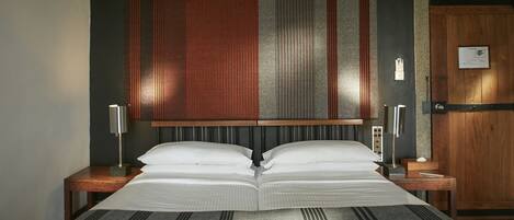 A double bed with a striped bedspread, two bedside tables with lamps, a wooden headboard, and a wooden door with a plaque.