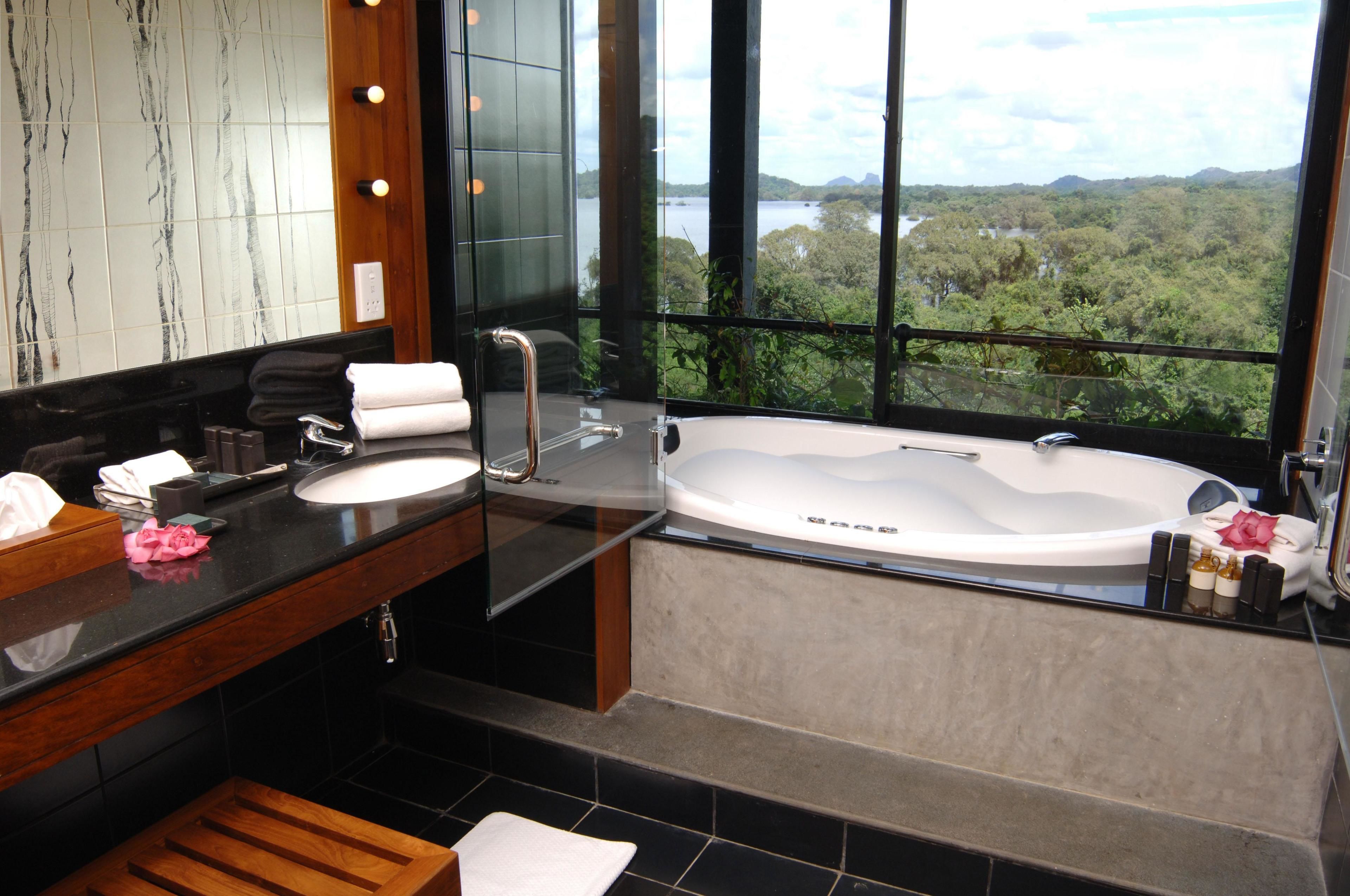 Un baño moderno con una bañera grande, un tocador con espejo y vistas a un paisaje boscoso a través de amplias ventanas.