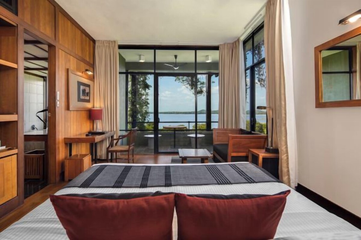 Habitación de hotel con una cama grande, un escritorio con silla, una mesita y un balcón con vista al agua.