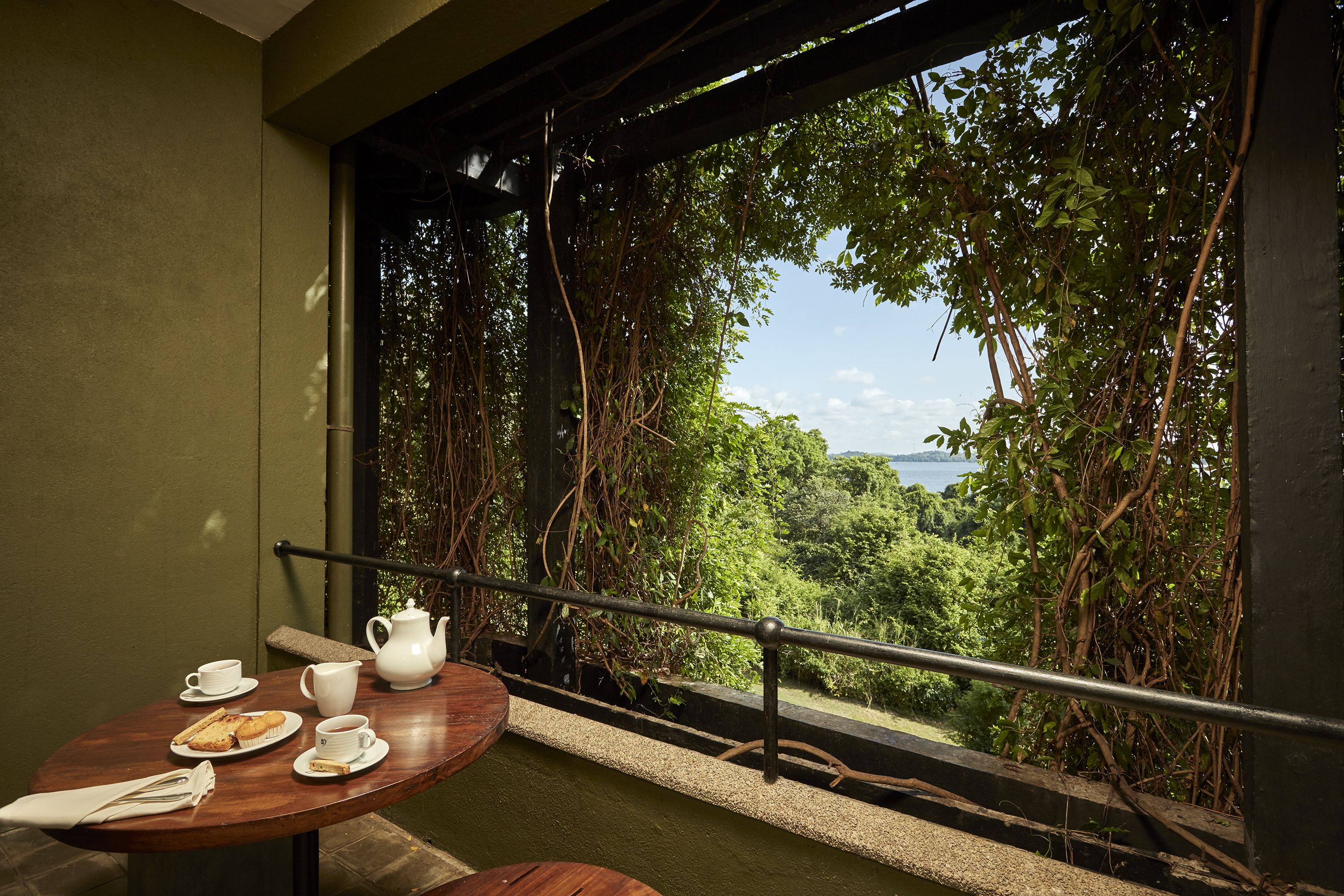 A balcony with a table set for tea, a view of lush greenery and a body of water.