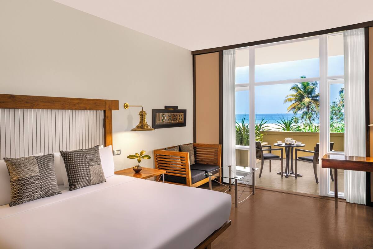 A hotel room with a large bed, a wooden chair, a small table, and a view of the ocean through large windows.