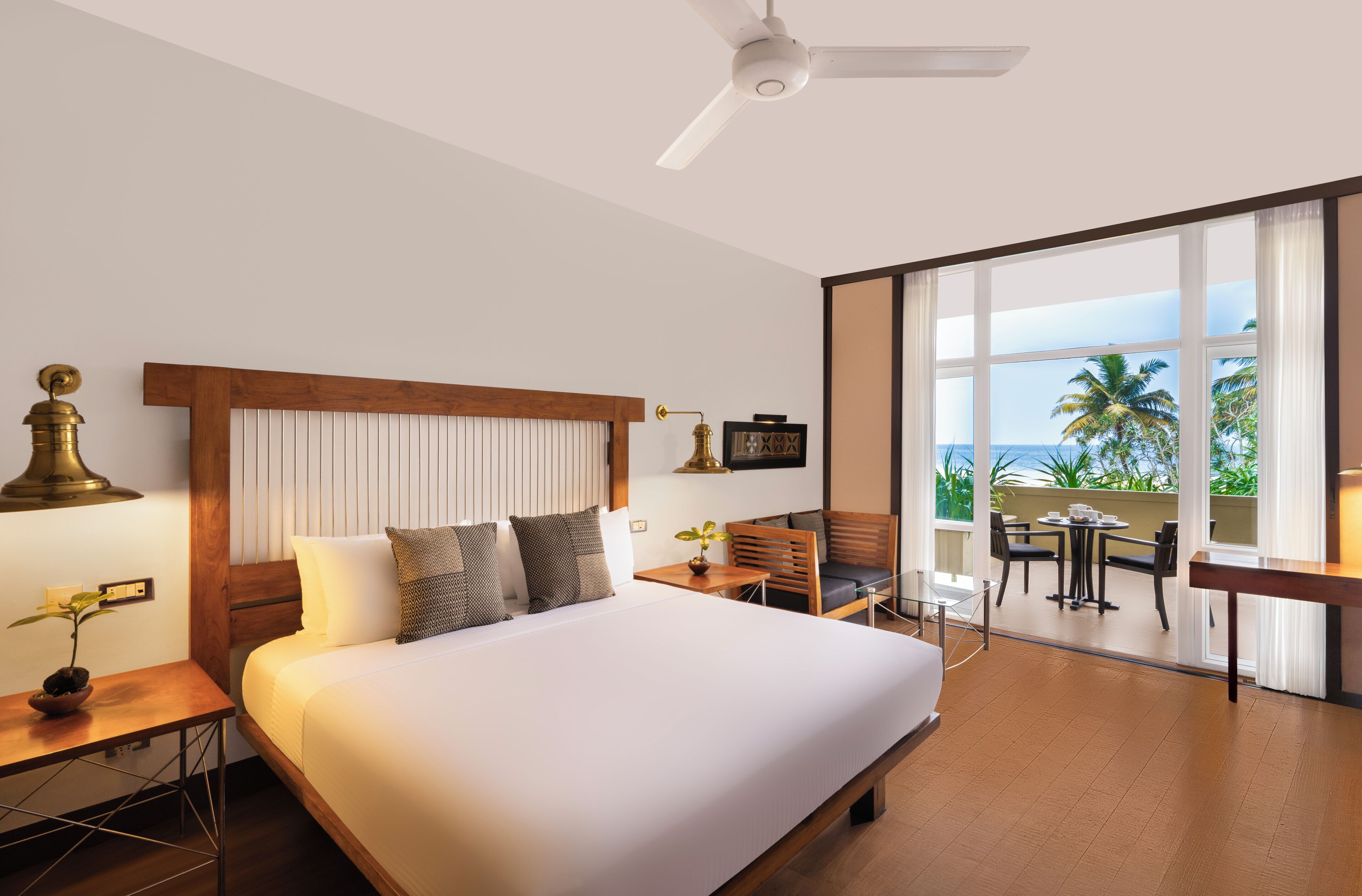 A hotel room with a large bed, a desk with a lamp, a small table with a plant, and a balcony with a view of the ocean.