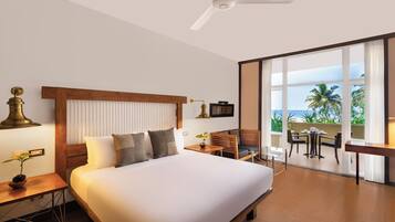 A hotel room with a large bed, a desk with a lamp, a small table with a plant, and a balcony with a view of the ocean.