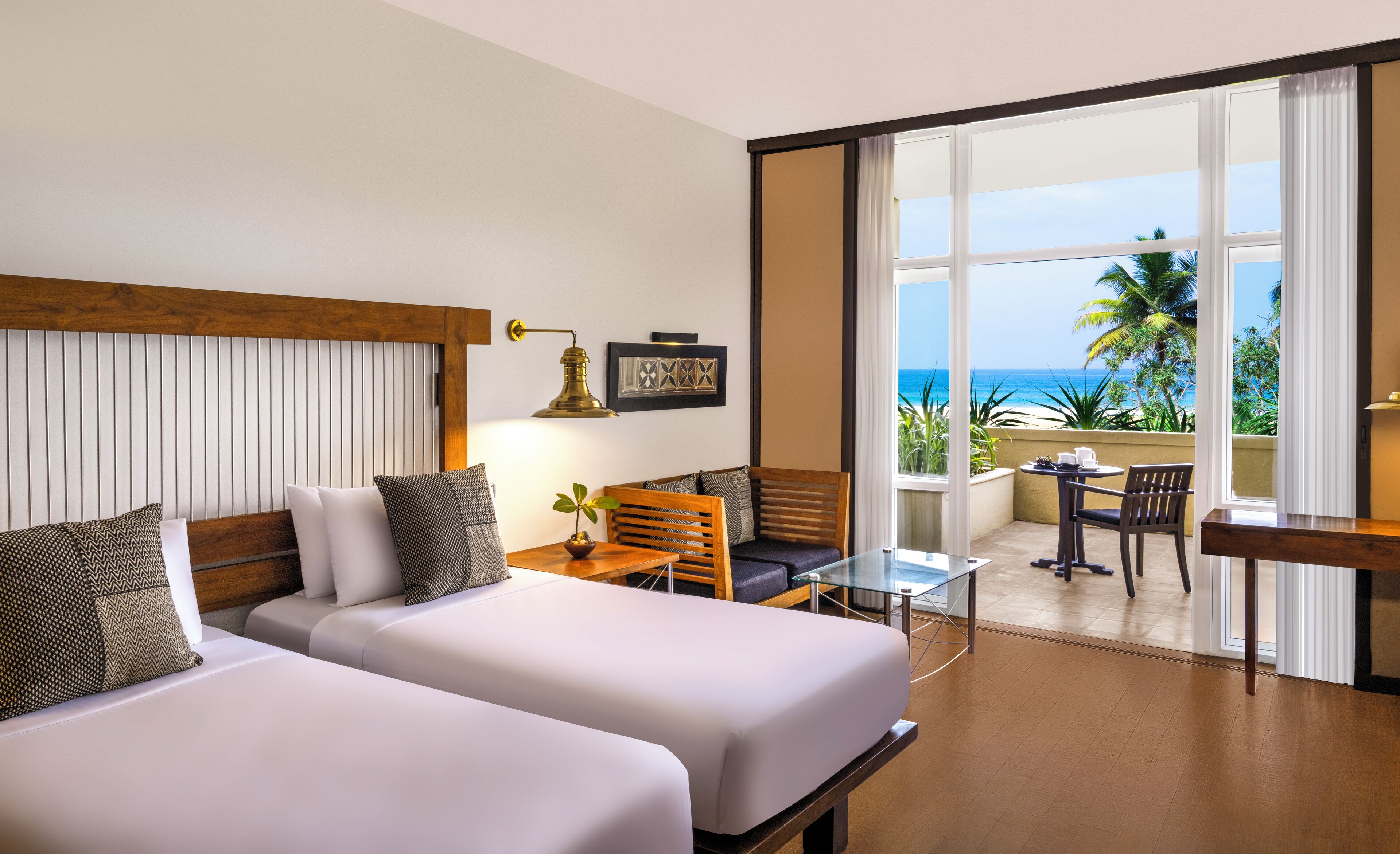 Habitación de hotel con dos camas, una mesita con sillas, un balcón con vista al océano y una ventana con cortinas.