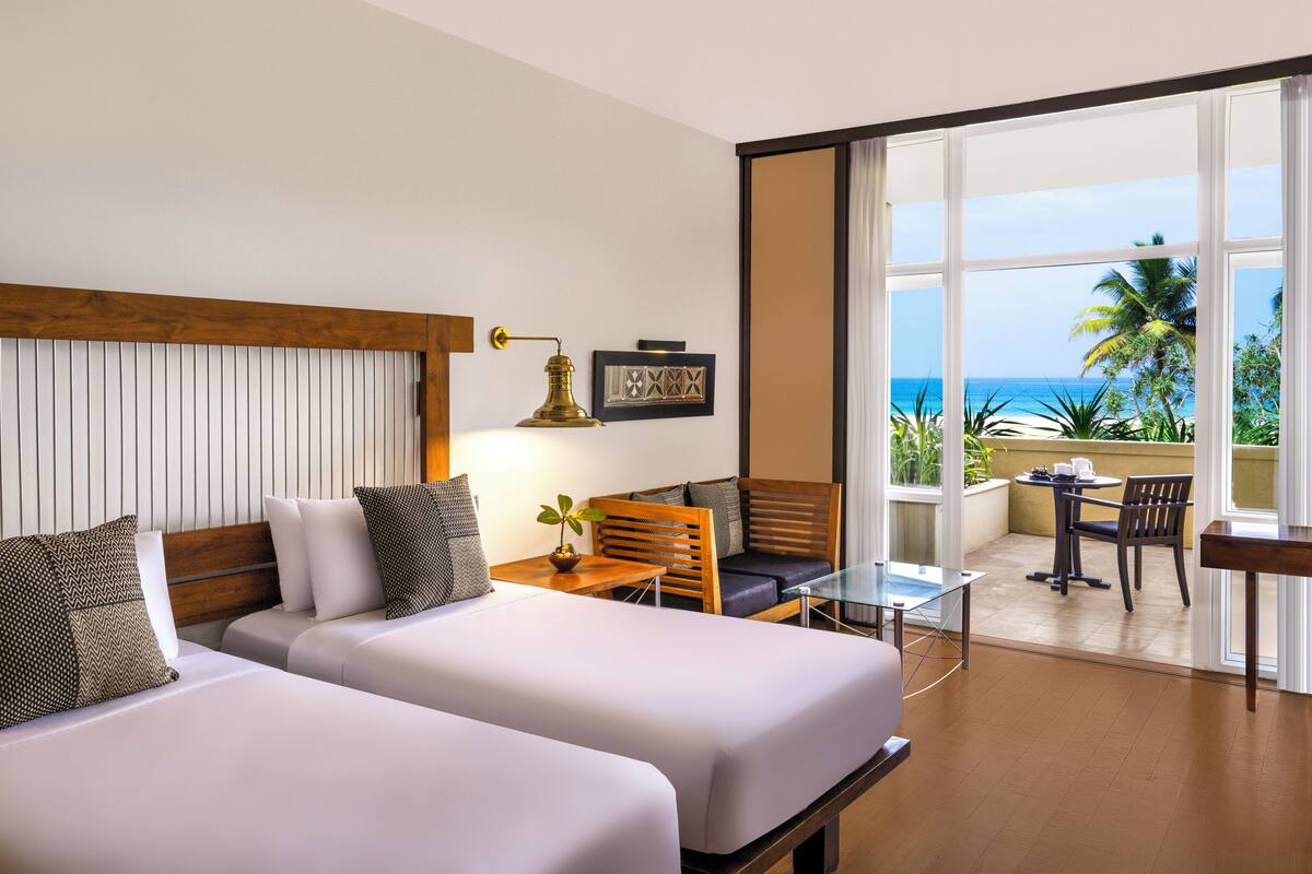 A hotel room with two beds, a small table with chairs, a balcony with a view of the ocean, and a window with curtains.